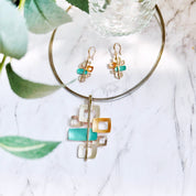 Matching Earrings and Necklace with a brown steel cable chain and a pendant with 6 interlocking squares plated in rose gold and painted in beige, gold, and teal enamel against a white marble background. Five squares contain square cut outs.