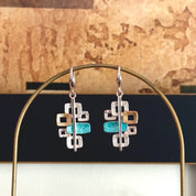 A pair of drop earrings with pendants with 6 interlocking squares plated in rose gold and painted in beige, gold, and teal enamel against a white background. Five squares contain square cut outs.