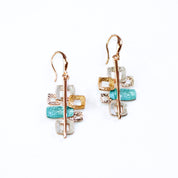 A pair of drop earrings with pendants with 6 interlocking squares plated in rose gold and painted in beige, gold, and teal enamel against a white background. Five squares contain square cut outs.