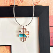 Necklace with a brown steel cable chain and a pendant with 6 interlocking squares plated in rose gold and painted in beige, gold, and teal enamel against a geometric abstract background. Five squares contain square cut outs.