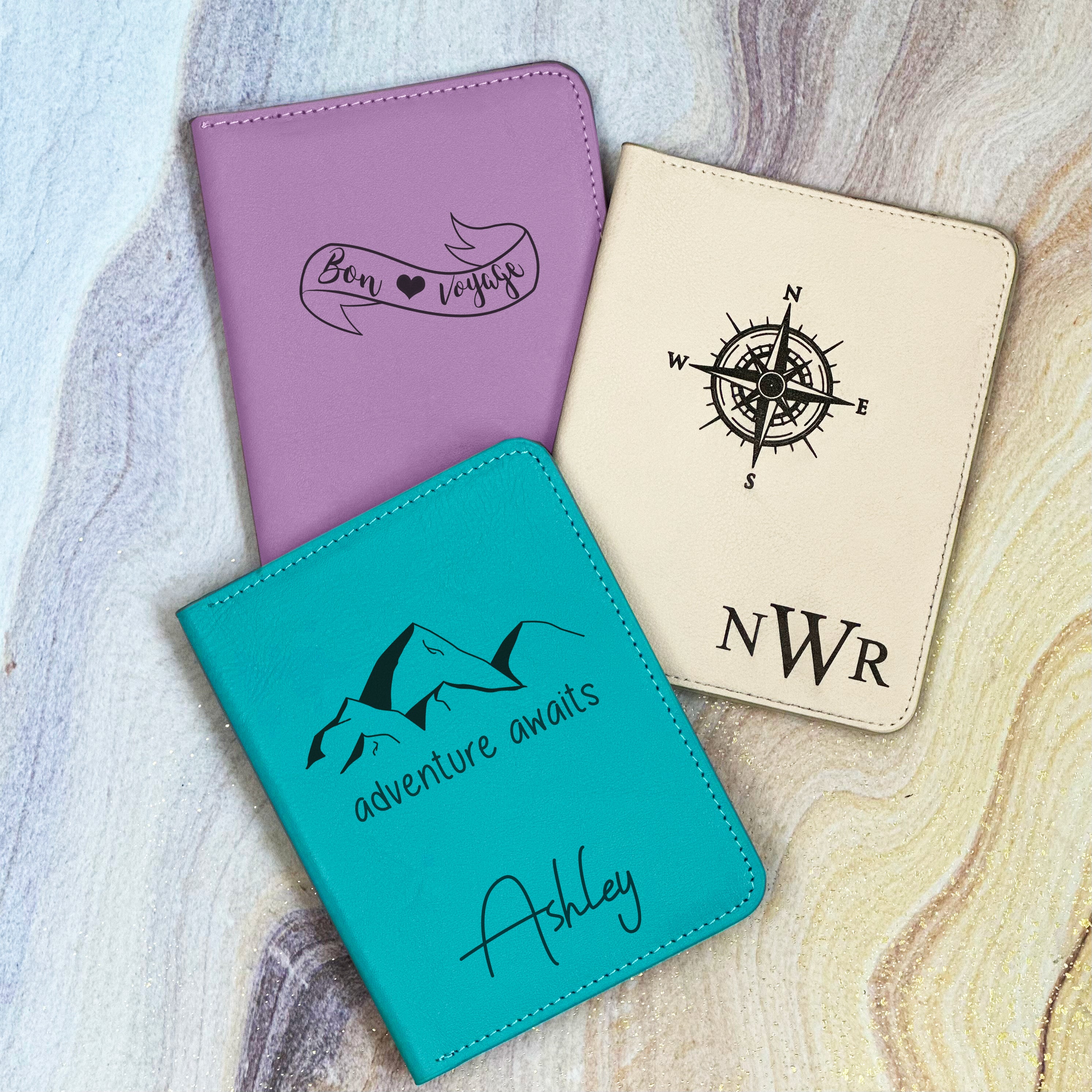 3 leather passport sleeves rest against a swirly background. A bright blue turquoise passport holder on the bottom of the image is engraved with a Mountain Range and the words Adventure Awaits underneath. The name Ashley engraved in Boulevard font sits at the bottom center of the cover. The white nude passport sleeve is monogrammed with the initials NWR in our Timeless monogram. A purple lavender passport holder is engraved with a banner outline and the words Bon Voyage with a heart in between the words.