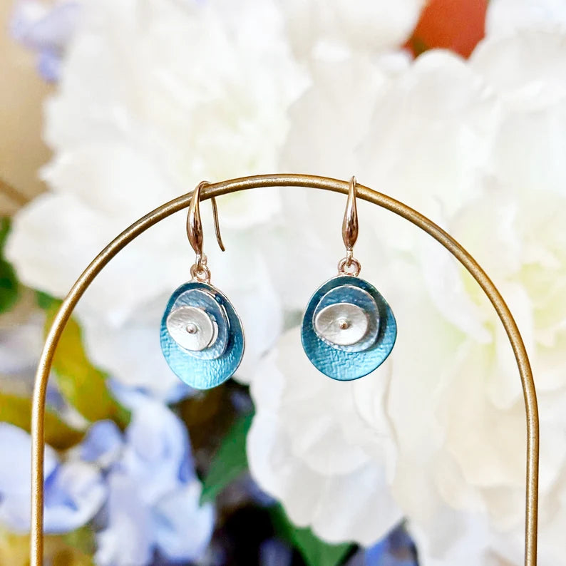 A pair of blue and silver boho abstract flower earrings hang off of a curved gold wire against a white and blue floral background. These beautiful earrings are lightweight and not too long, making them essential for day to day wear. Dress them up with the matching necklace for a more glamorous effect - why not go big? Personalized upon request, these earrings can be made with your 3 favorite colors. Anything is possible!