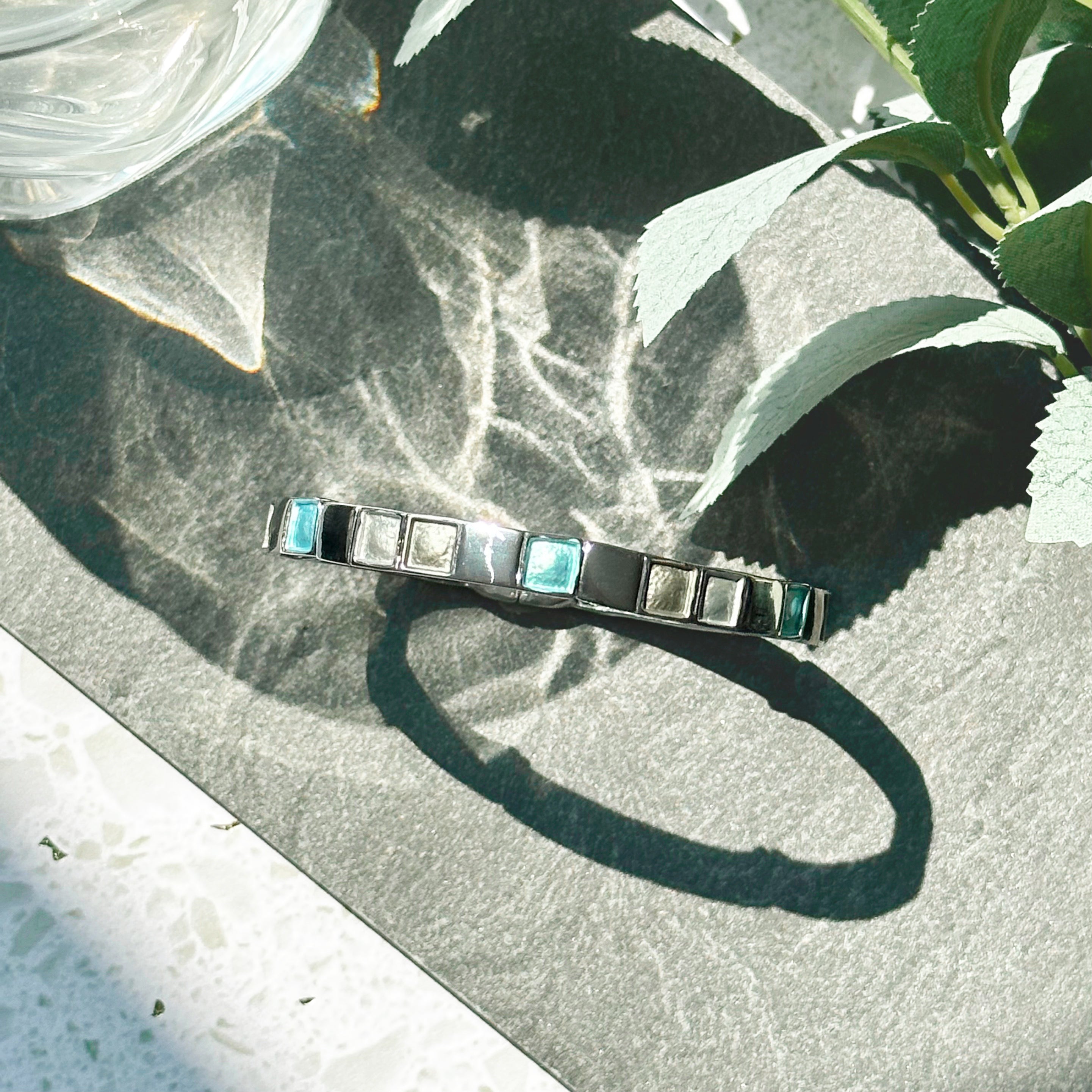 Rhodium plated cuff bracelet with a magnetic closure with square indents painted in silver, gray, and teal enamel is placed upright on a black stone tile with shadows cast by a glass and a leafy plant