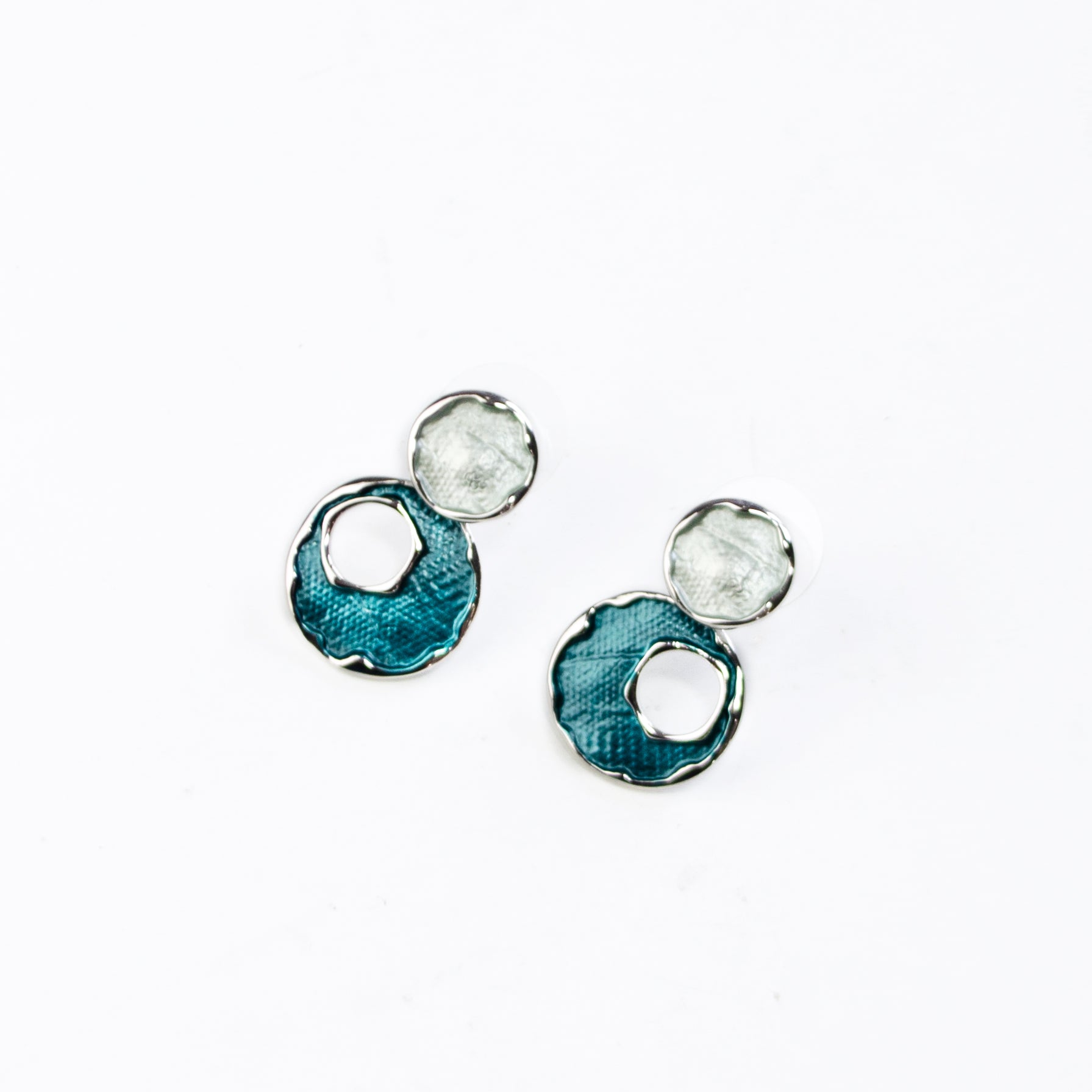 A pair of rhodium plated stud earrings made with two circles, the top painted with silver enamel, and the bottom larger circle painted in blue enamel with an off-center circle cutout rest on a marble tile with leafy plants on a white stone background