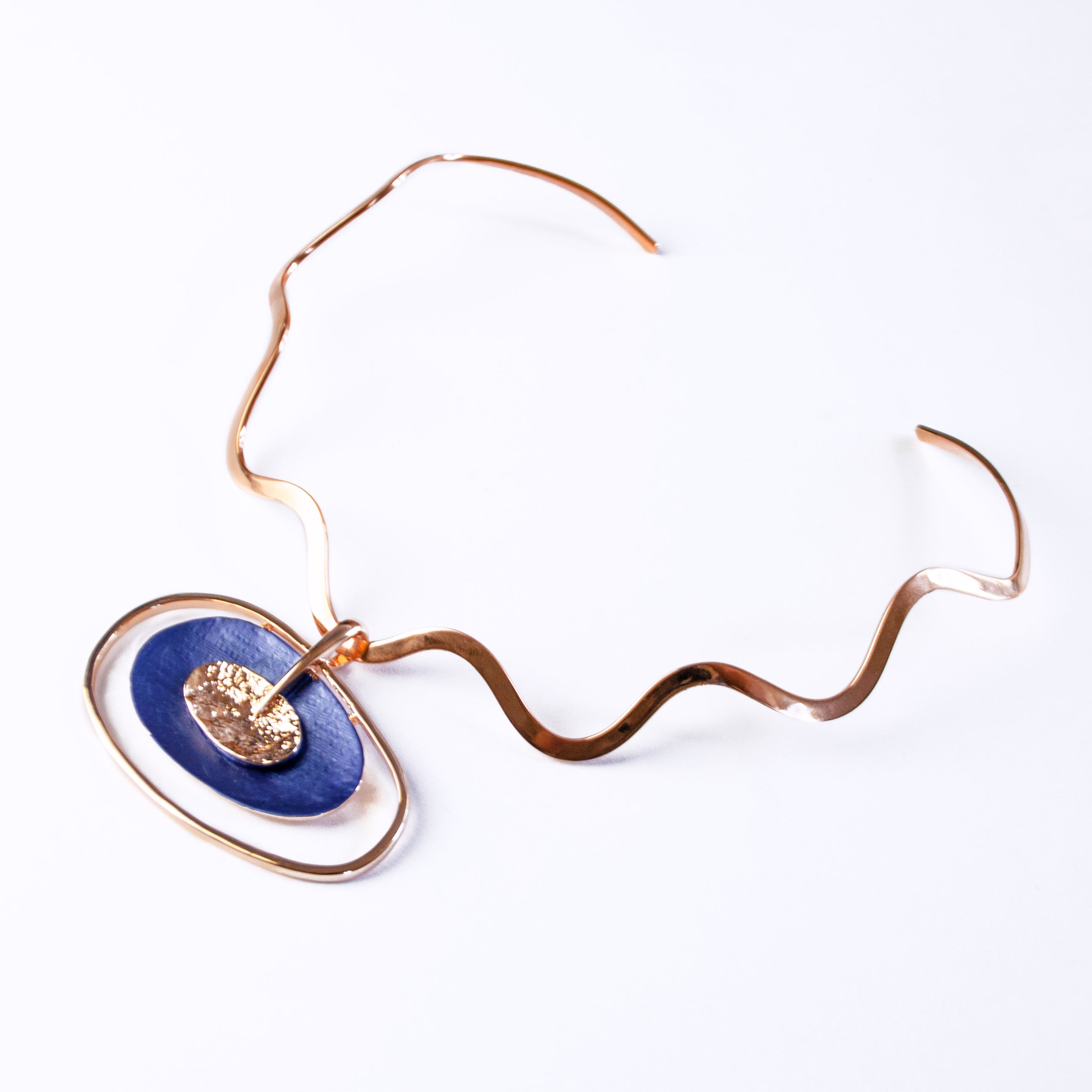 rose gold plated Open Oval Pendant with two textured overlapping oval disks in the center, painted in blue enamel and sitting on a copper wavy neck ring against a white background