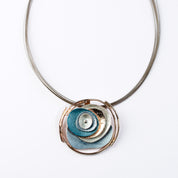 Adjustable Necklace with Brown Steel Wire chain and a round pendant made of 6 rose gold plated overlapping abstract textured discs painted in teal, blue, and gray enamel against a white background