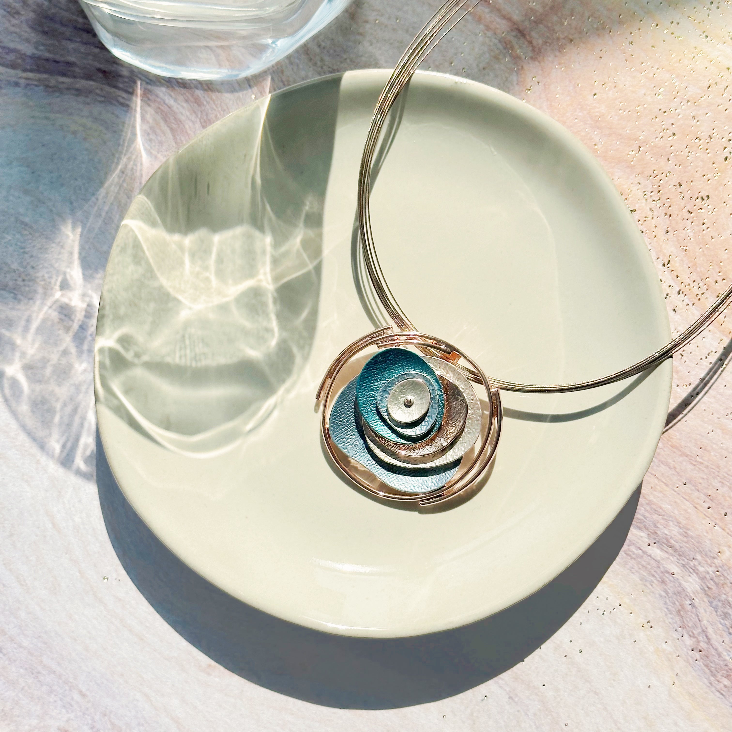 Adjustable Necklace with Brown Steel Wire chain and a round pendant made of 6 rose gold plated overlapping abstract textured discs painted in teal, blue, and gray enamel rests on a white plate on a purple and peach stone background