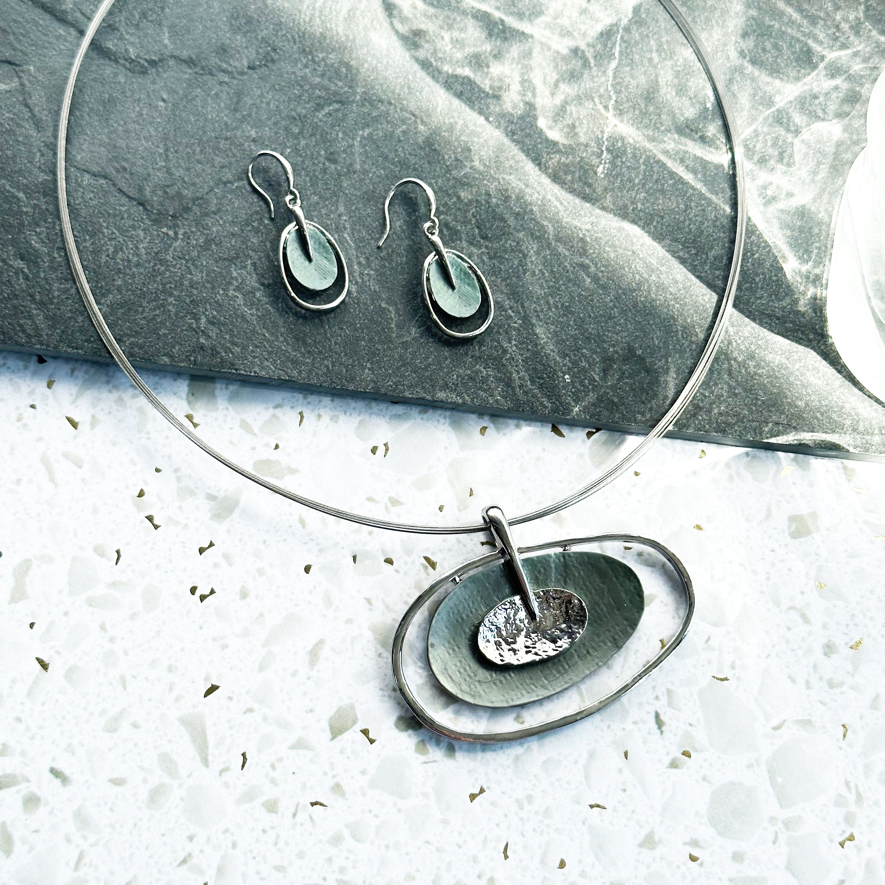 A pair of rhodium plated open oval drop earrings with hook backings and a textured oval disc at the center painted in green enamel rest on a textured gray stone tile on a white stone background with a matching necklace on a rhodium plated choker