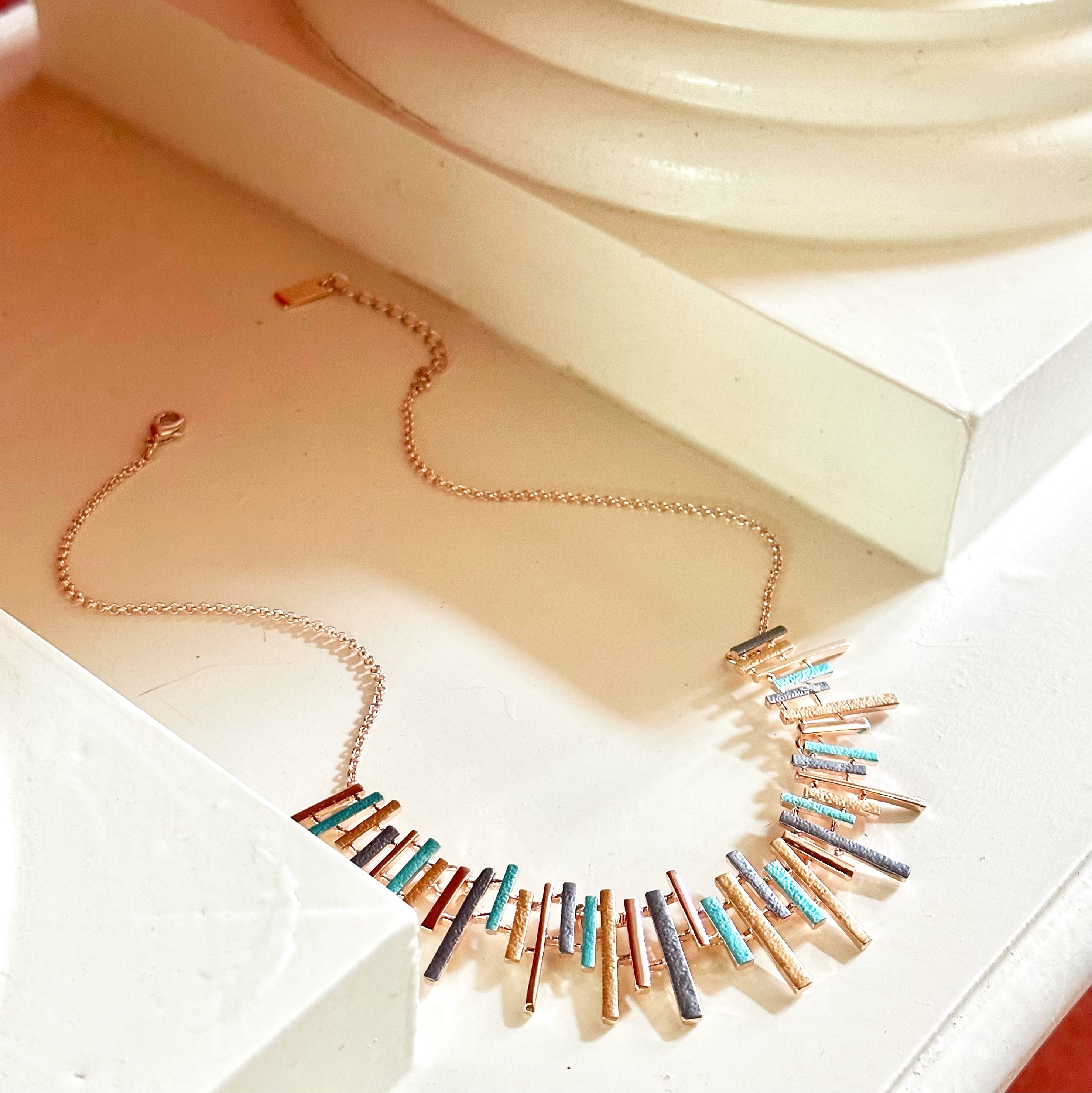 Adjustable Rose Gold Plated Necklace with a cable chain and connected pendants with vertical bars of varying lengths painted in teal, gray, and gold enamel sitting in between the bottom of two columns