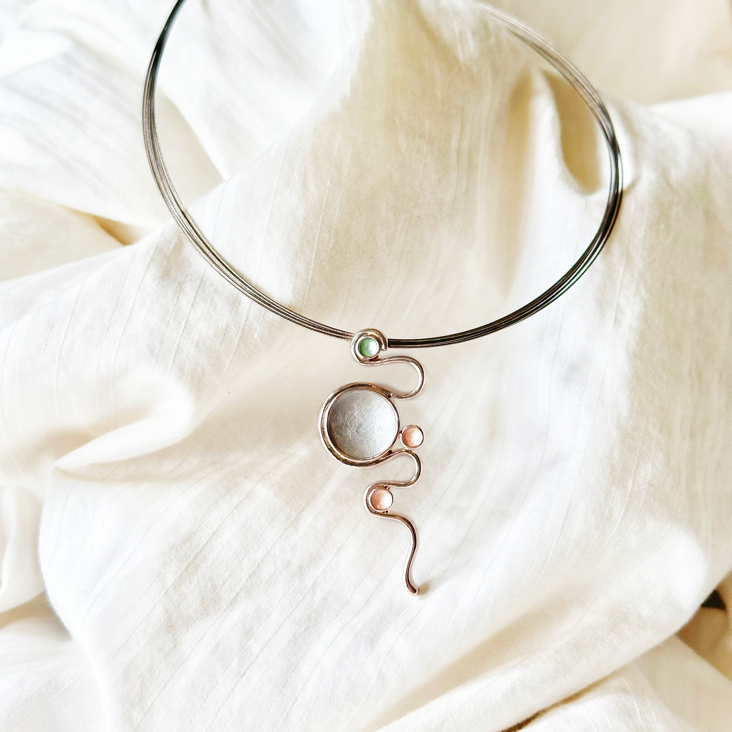 Adjustable Necklace with a brown steel wire chain and a rose gold plated pendant with a serpent snaking around three small circles and one large circle individually painted in green, gray, and gold enamel against a beige cloth background