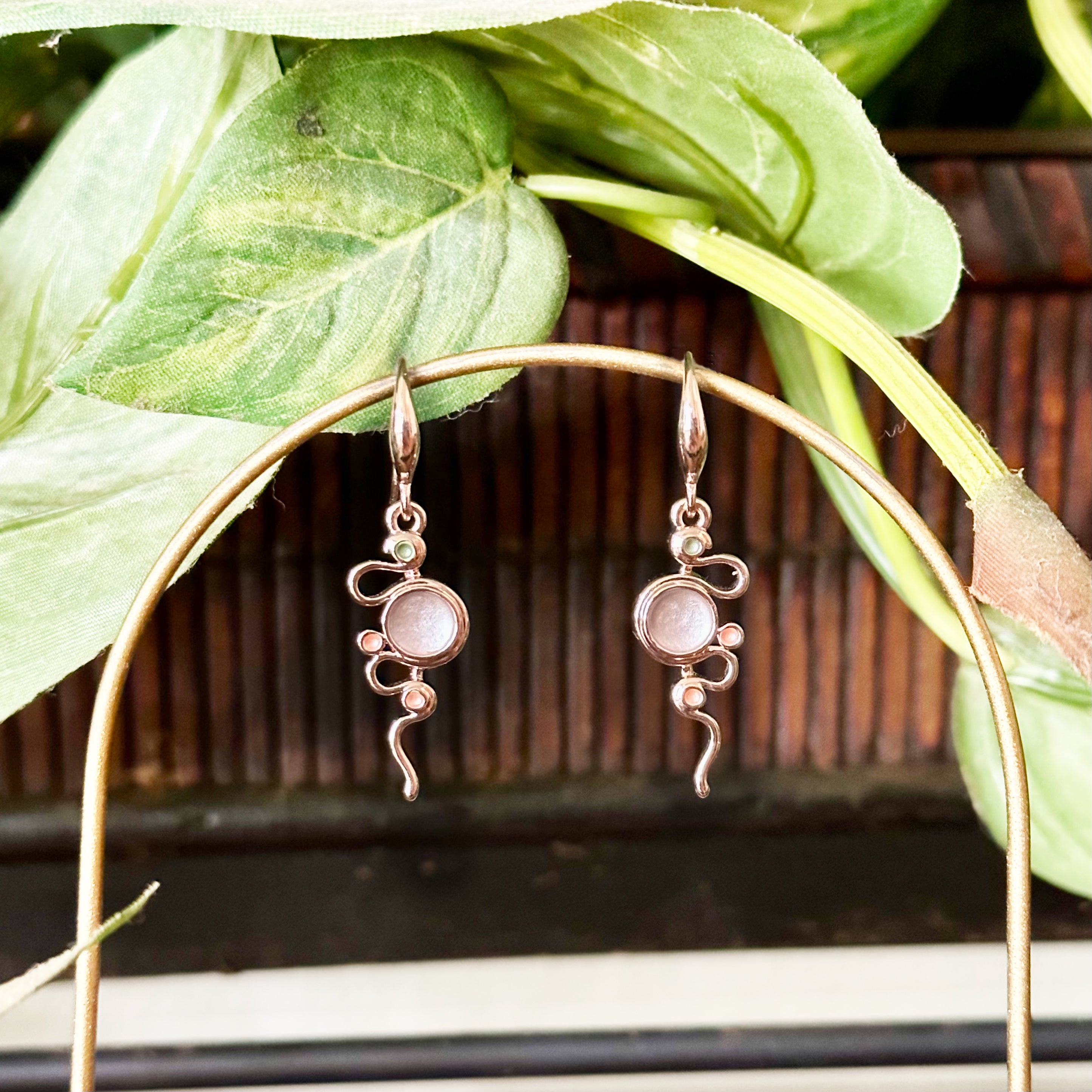 Rose Gold Plated Dangle Earrings against a plant background with a hook back and a thin rose gold serpent snaking around three small circles and one large circle each painted in green, gray, and gold enamel