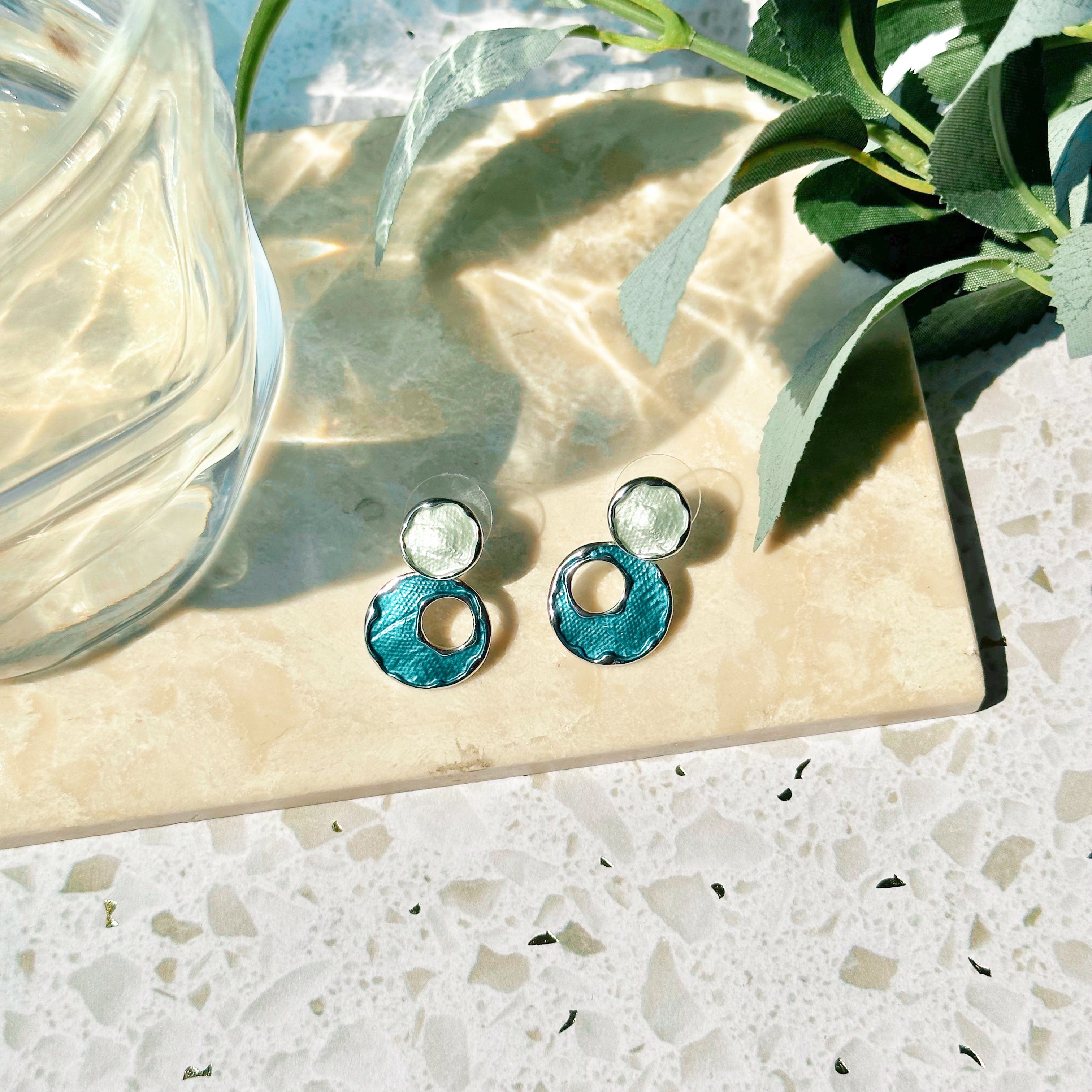 A pair of rhodium plated stud earrings made with two circles, the top painted with silver enamel, and the bottom larger circle painted in blue enamel with an off-center circle cutout rest on a marble tile with leafy plants on a white stone background