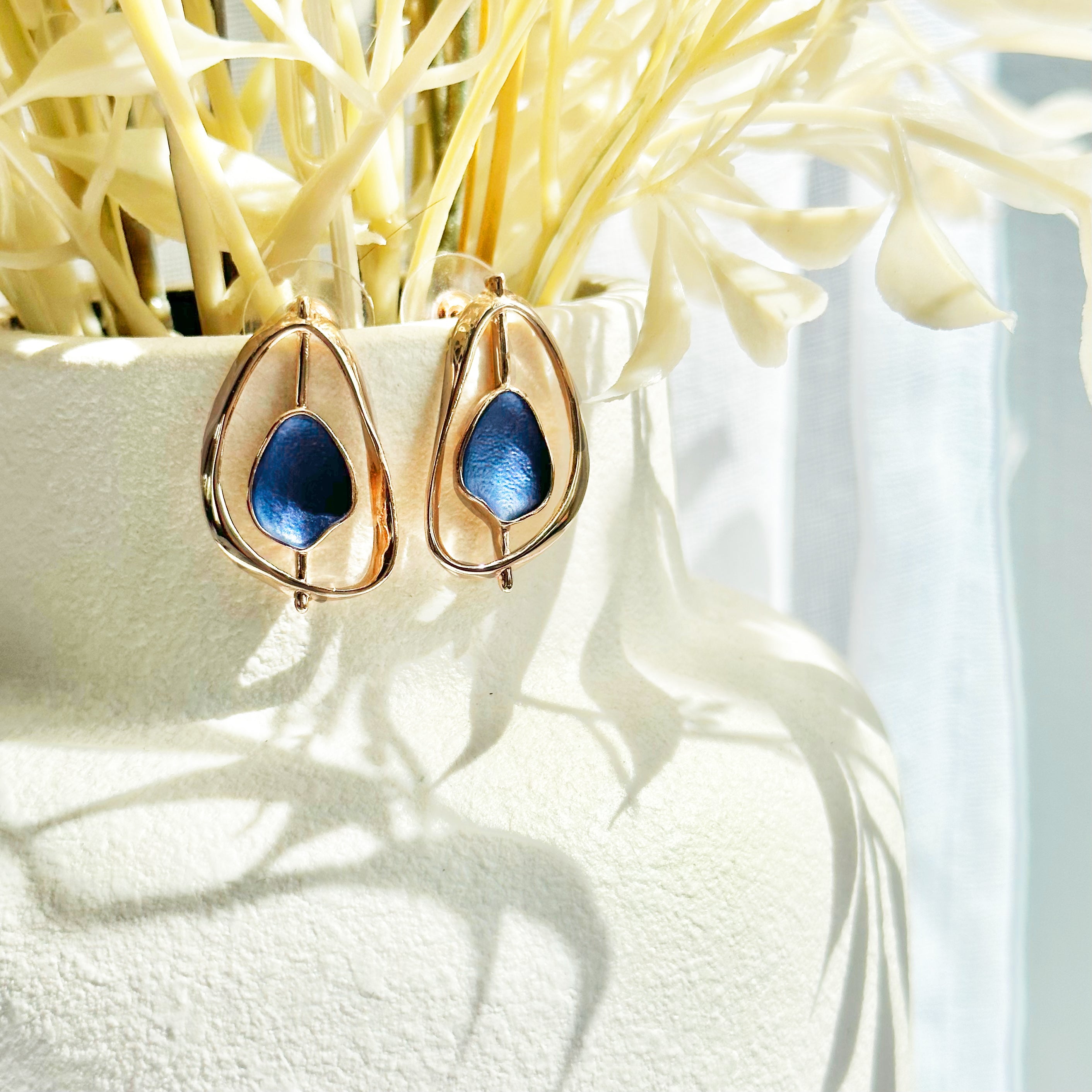 Open Pear shape rose gold stud earrings with a floating textured disc center painted in blue enamel sit on the top of a white vase with beige plant stems coming out the top casting shadows on the vase.