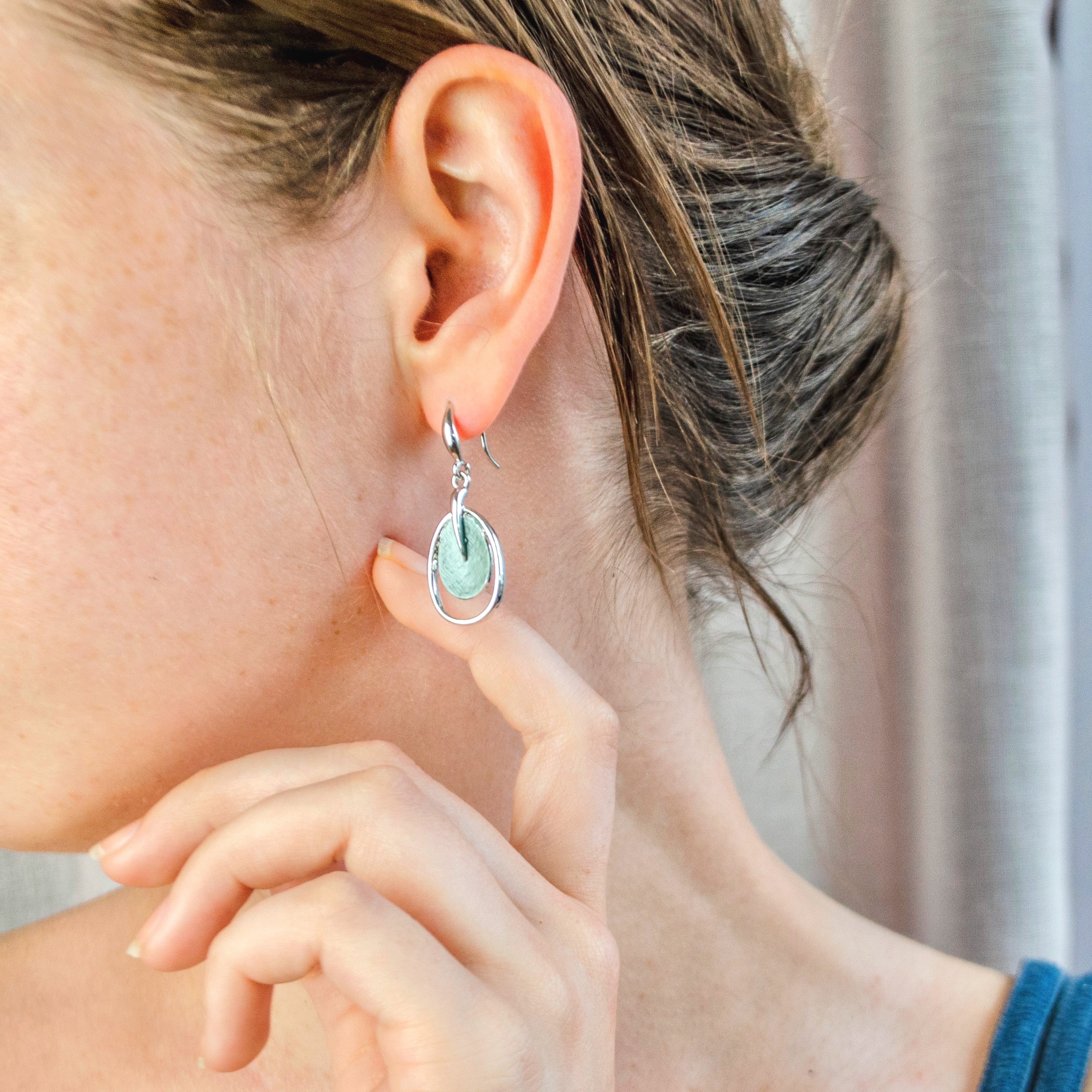 A pair of rhodium plated open oval drop earrings with hook backings and a textured oval disc at the center painted in green enamel are worn by a model with her head turned to the side and her hand by her jawline