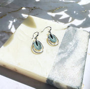 A pair of rhodium plated open oval drop earrings with hook backings and a textured oval disc at the center painted in green enamel rest on a stone white and gray color block coaster underneath a plant casting shadows