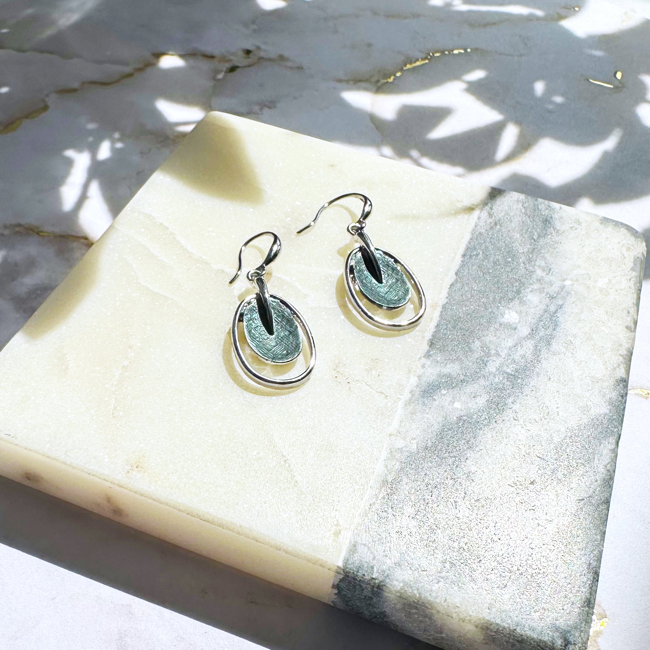 A pair of rhodium plated open oval drop earrings with hook backings and a textured oval disc at the center painted in green enamel rest on a stone white and gray color block coaster underneath a plant casting shadows