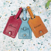 3 luggage tags in raspberry, silver gray and honey sit on a multicolored background. Engraved name bag tag, engraved monogram bag tag, engraved date bag tag, luggage id tag with date engraving, name engraving travel kit accessories for overseas trip