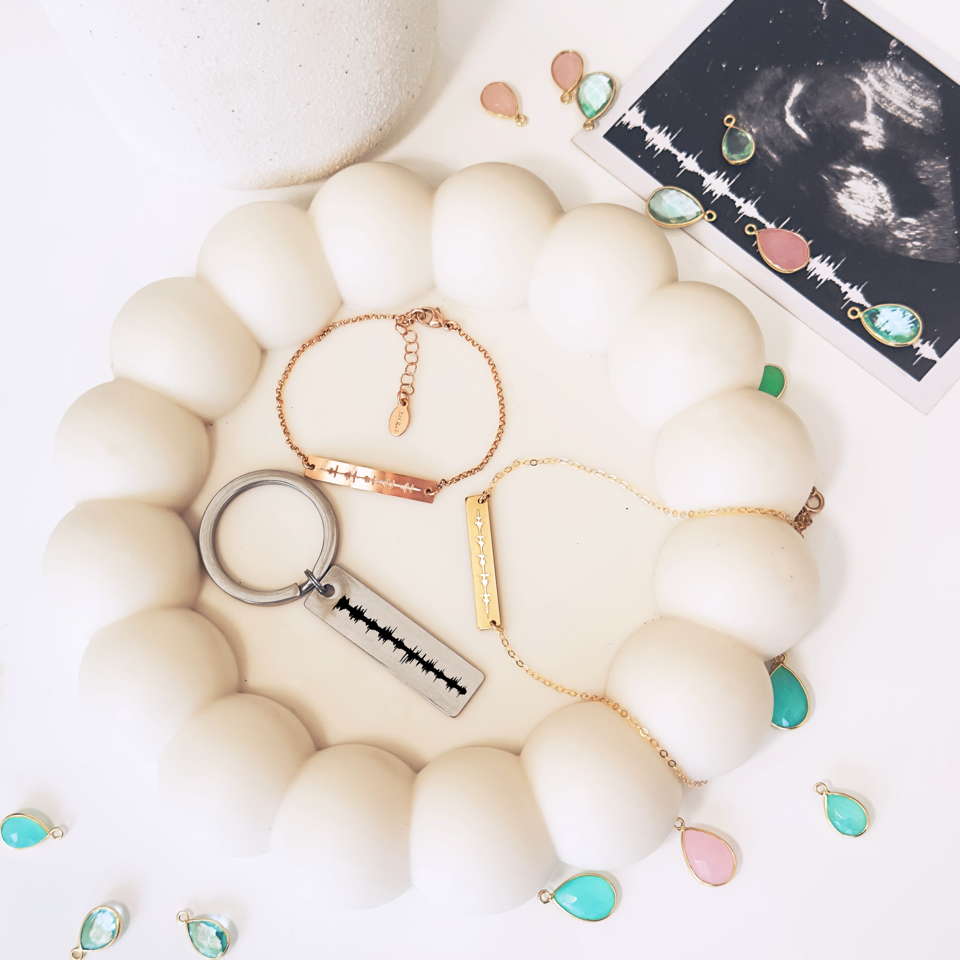 An image of a heartbeat keychain, necklace and bracelet sitting in a rounded dish with pink and blue crystals scattered around. Each item in our heartbeat collection is designed to honor the unique and individual heartbeat of your newborn baby