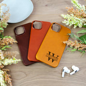 Three phone cases in dark brown espresso, vibrant cinnamon, and classic honey colors are laid overlapping against a wood background. There are flowers on either side and a pair of Airpods below the honey phone case. There is a white dish in the upper left corner above the espresso colored phone case. Phone case gift for partner,  best unexpected gift for husband, gift for men, personalized gift for men, engraved men's phone case iPhone 15, new iPhone case, engraved phone case for him or her