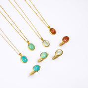 Four Gold Necklaces with oval cabochon pendants with matching Gold rings below. From Left to Right: Blue Amazonite on a cable chain, mint green Chrysoprase on a satellite chain, Mother of Pearl on a cable chain, and Sunstone on a satellite chain