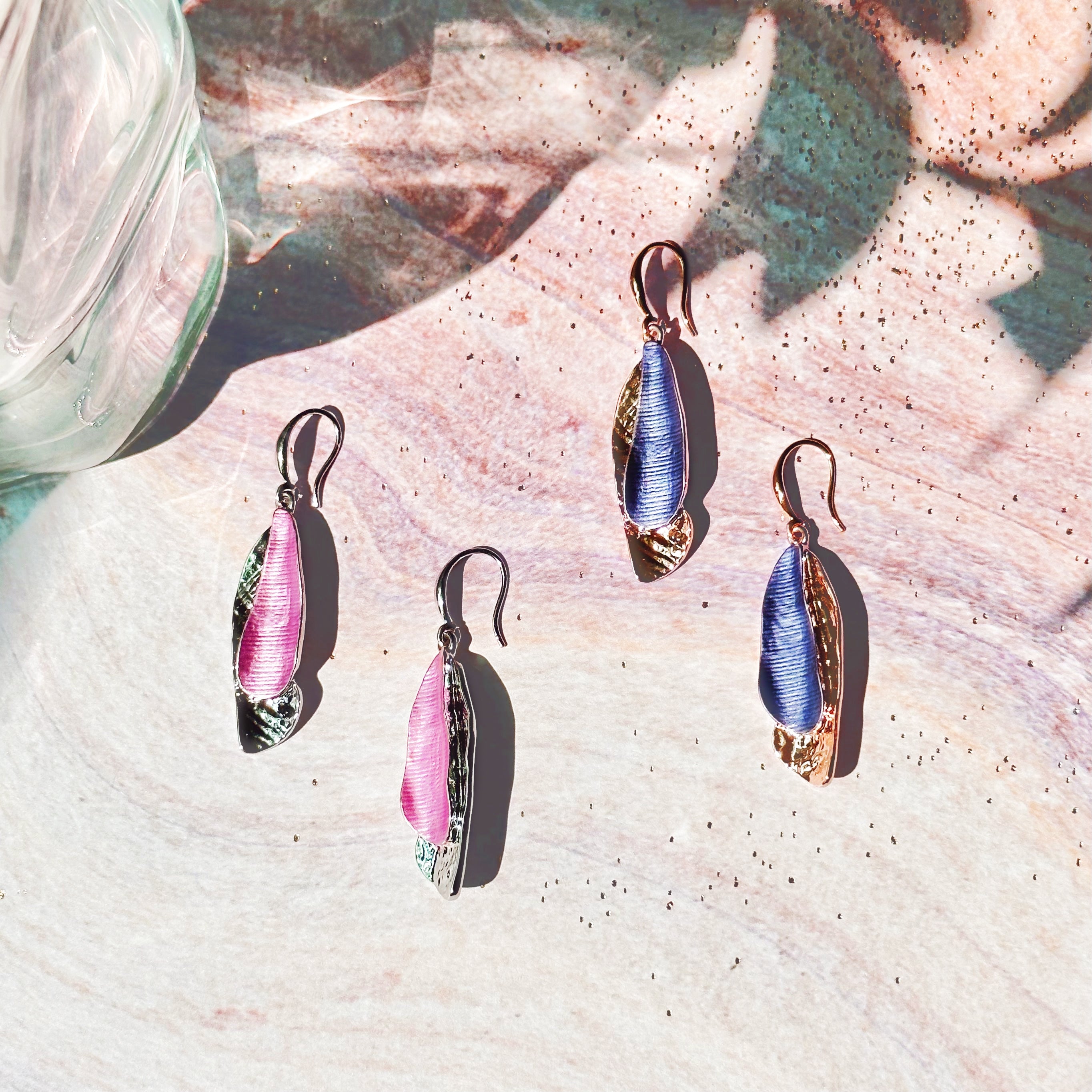 Two sets of Hook Back Drop Earrings with pendants made of two overlapping textured organic shaped bars with the bottom bar plated in rhodium or rose gold and the top painted in pink or purple enamel sit against a pink swirling stone background