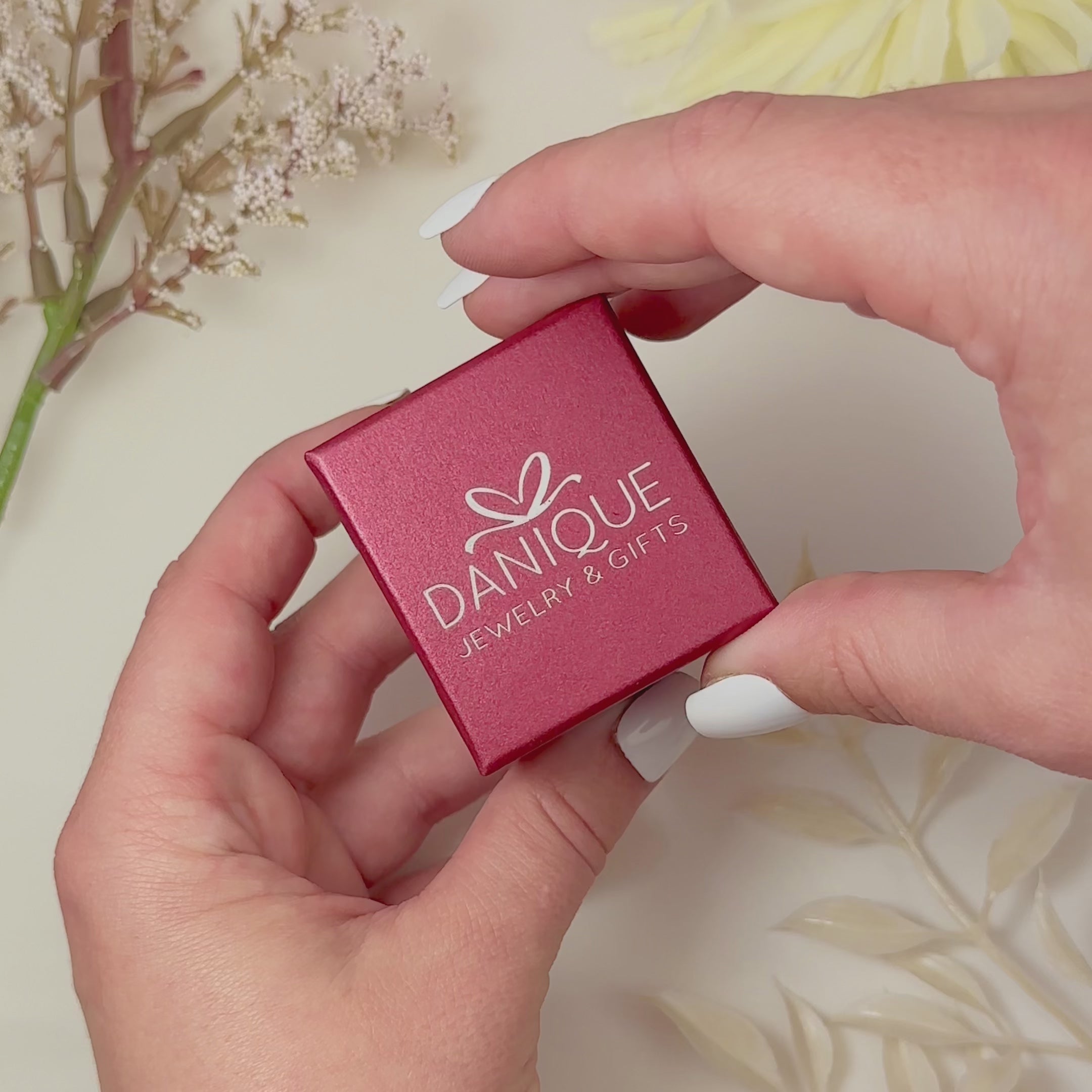 2 hands hold and open a red Danique Jewelry ring box, revealing a set of gold rings with an oval pink chalcedony ring and 3 tiny round rings in blue topaz, pink chalcedony and green onyx. White flowers are in the corner of the image. 2 hands show a peridot oval ring revealing an open back and thin flat band. A tiny ruby ring is shown with an open back and rounded band. A panover shows a set of silver, rose gold, and gold rings in open boxes A hand in the corner wears a gold stack of mixed birthstone rings. 