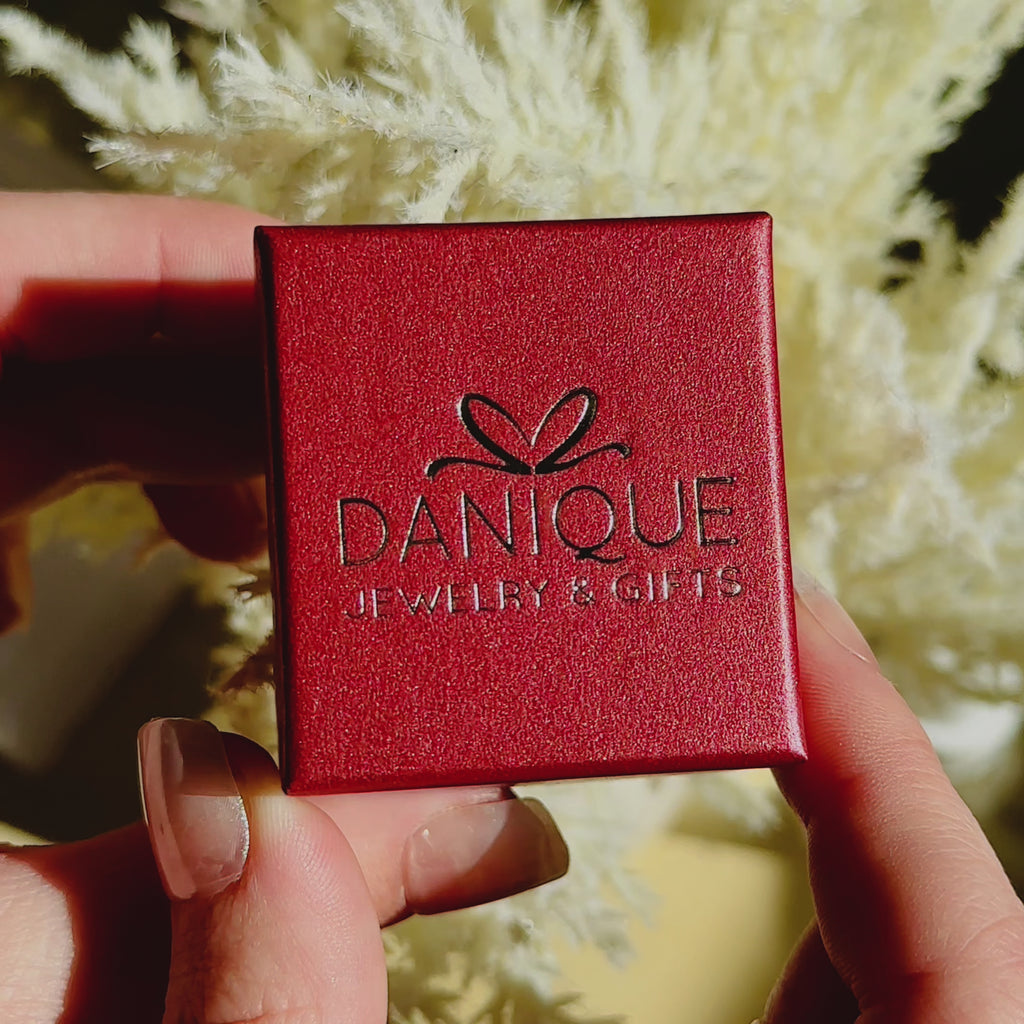 A person opens a red Danique Jewelry ring gift box to reveal a gold vermeil engraved oval birth flower ring. A person wears a silver engraved birth flower ring with a stackable citrine diamond shaped ring against a tree. A hand holds a silver engraved birth flower ring against a blue sheet background. An up close shot of a person's hand wearing a sterling silver ring. 