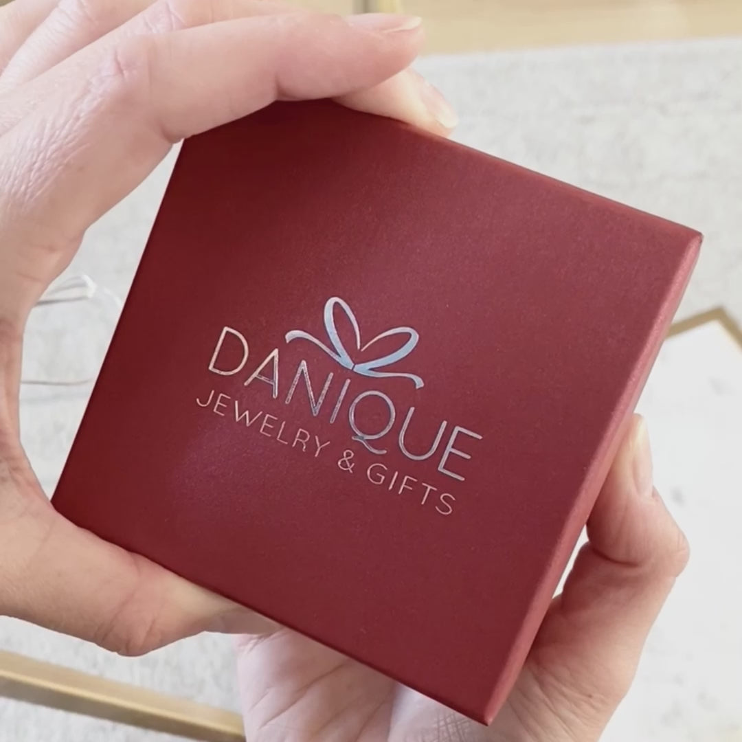 Hands open a red square Danique Jewelry box to reveal a gold cascade necklace with oval name tags and green onyx, kyanite quartz and ruby quartz birthstones in half prong half bezel settings. A person with shoulder length brown hair wears a gold cascade necklace with white and pink stones and oval name tags. A child gifts a red Danique Jewelry box to her mother. A group of silver and gold cascade necklaces dangle and sparkle next to each other. The child and mother show a gold necklace with initial charms.