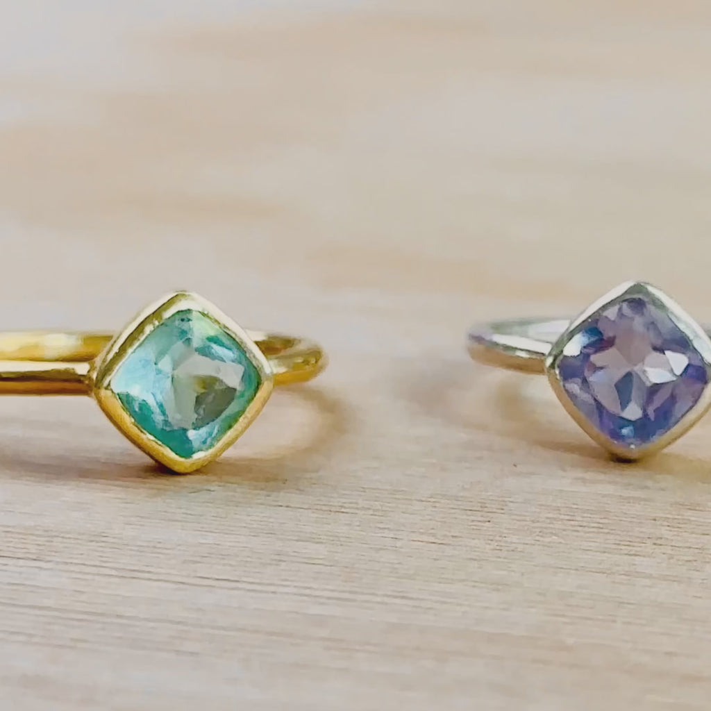 The camera pans to the right to reveal a variety of silver and gold diamond shaped rings. Two hands show a blue topaz december birthstone diamond shaped ring up close. A hand wears a rainbow moonstone june birthstone ring. A hand with long purple nails wears a blue kyanite september birthstone and a light purple alexandrite quartz june birthstone ring set. Two hands show an open red Danique Jewelry ring box with a kyanite and alexandrite quartz.