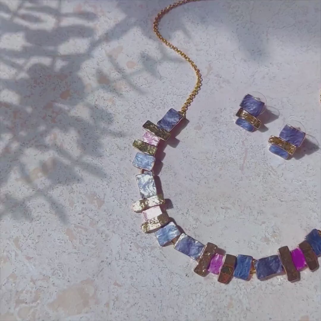 This video features an array of creations from our Geometric Bohemian Collection. The first panover shows a geometric squares set of earrings and a necklace in pink and purple with a rose gold finish. A hand shows a set of geometric layered square earrings and a matching necklace. A person wears a geometric squares bangle bracelet in silver with teal and blue. A panover shows a sunray linear necklace in rose gold with yellow, blue and silver accent colors on the sun's "rays". Custom colors are available. 