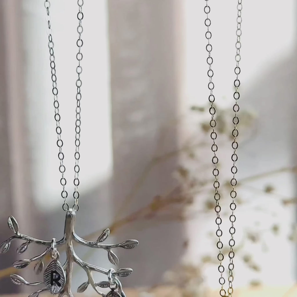 The camera pans down to reveal a gold and a silver upside down family tree necklace with dangling hand stamped leaf initial charms. A person runs their thumb over the pendant to show the movement and size. A person in a black shirt wears a gold tree necklace.  A person opens a red danique jewelry gift box to show a silver leaf necklace inside. This is the perfect gift for mom or grandma for christmas, birthdays, anniversaries, or any special gift giving occasion.