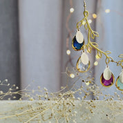 The camera pans to the right, revealing a gold double branch necklace with birthstones and engraved name charms. A person in a white v neck shirt wears the branch necklace and turns to the right. Two hands show an up close shot of the double branch necklace. Two hands hold a silver branch necklace in an open red Danique Jewelry box. 