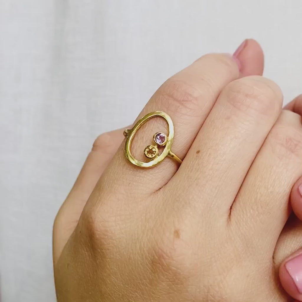 A person wears a 2-stone 3mm amethyst and citrine open oval ring. This delicate egg shaped piece can be set with your choice of birthstone, with gemstone options for every month, plus alternative options if you want something a little different. Rings are available in gold, rose gold, or sterling silver and are hand crafted to your unique style. Birthstones available for January, February, March, April, May, June, July, August, September, October, November, December. 