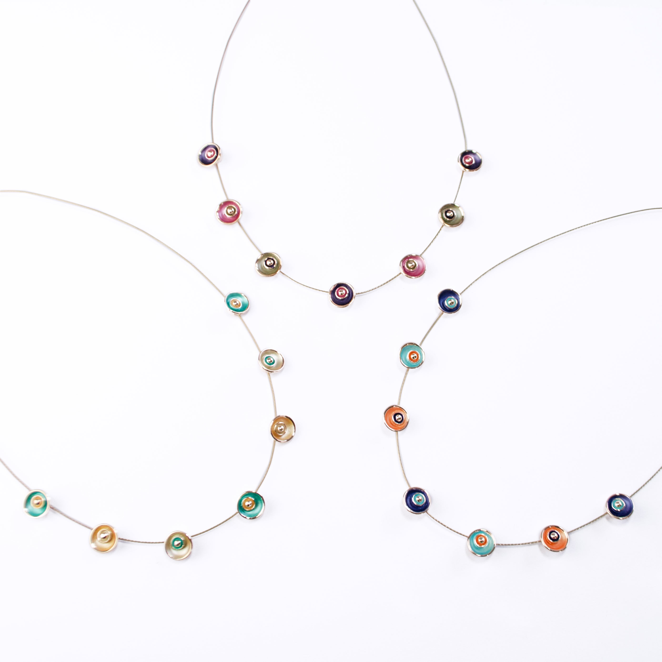 Three adjustable Choker necklaces on a white background with brown steel cable chains, each with seven rose gold plated circular flower charms, one painted in teal, gold, and beige, one in blue, teal and orange, and one in purple, pink, and gray