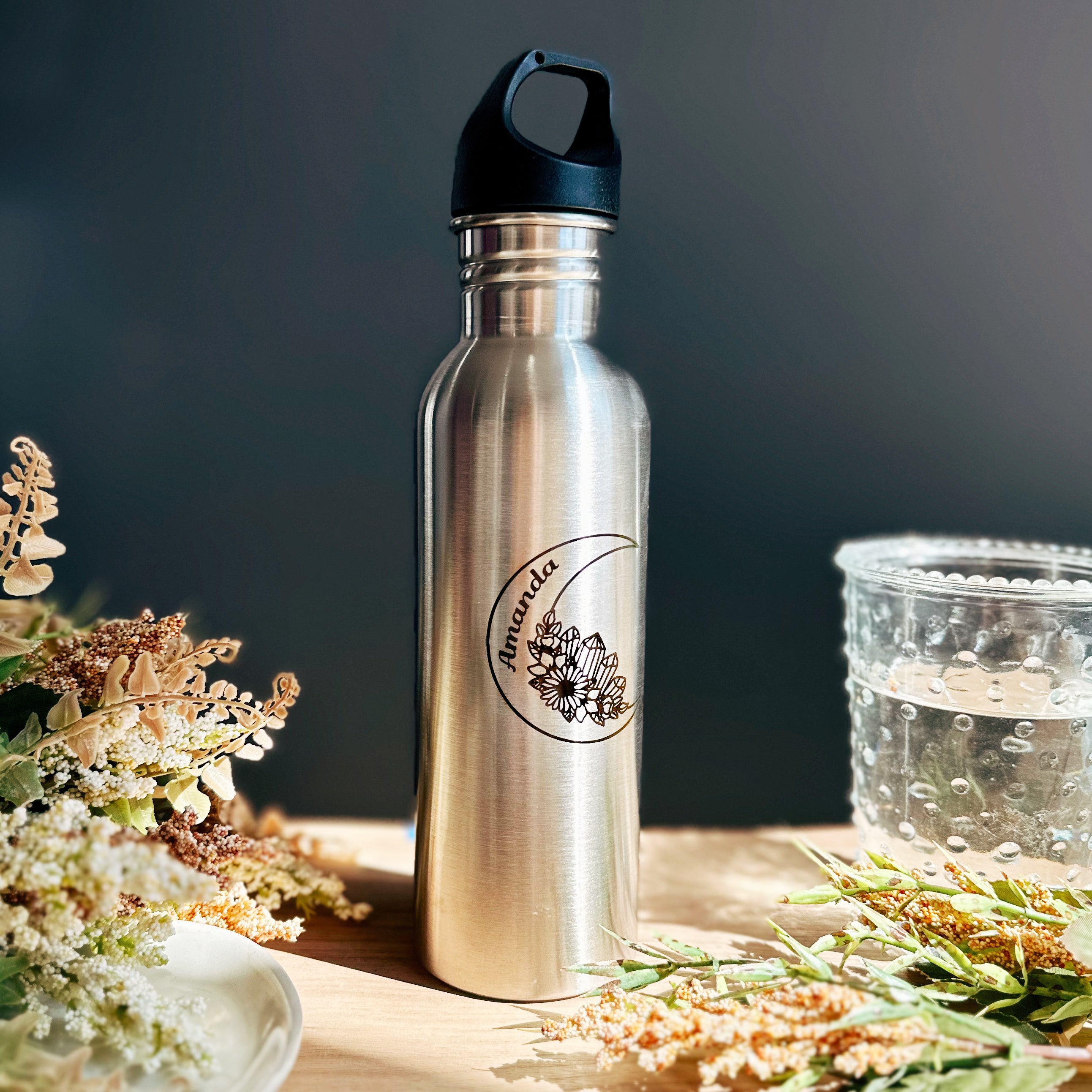 A silver stainless steel water bottle engraved with a crescent moon shape with flowers and crystals and the name Amanda in the moon sits on a wood table with flowers and a glass. Sunlight shows the clear engraving on the high quality steel bottle.