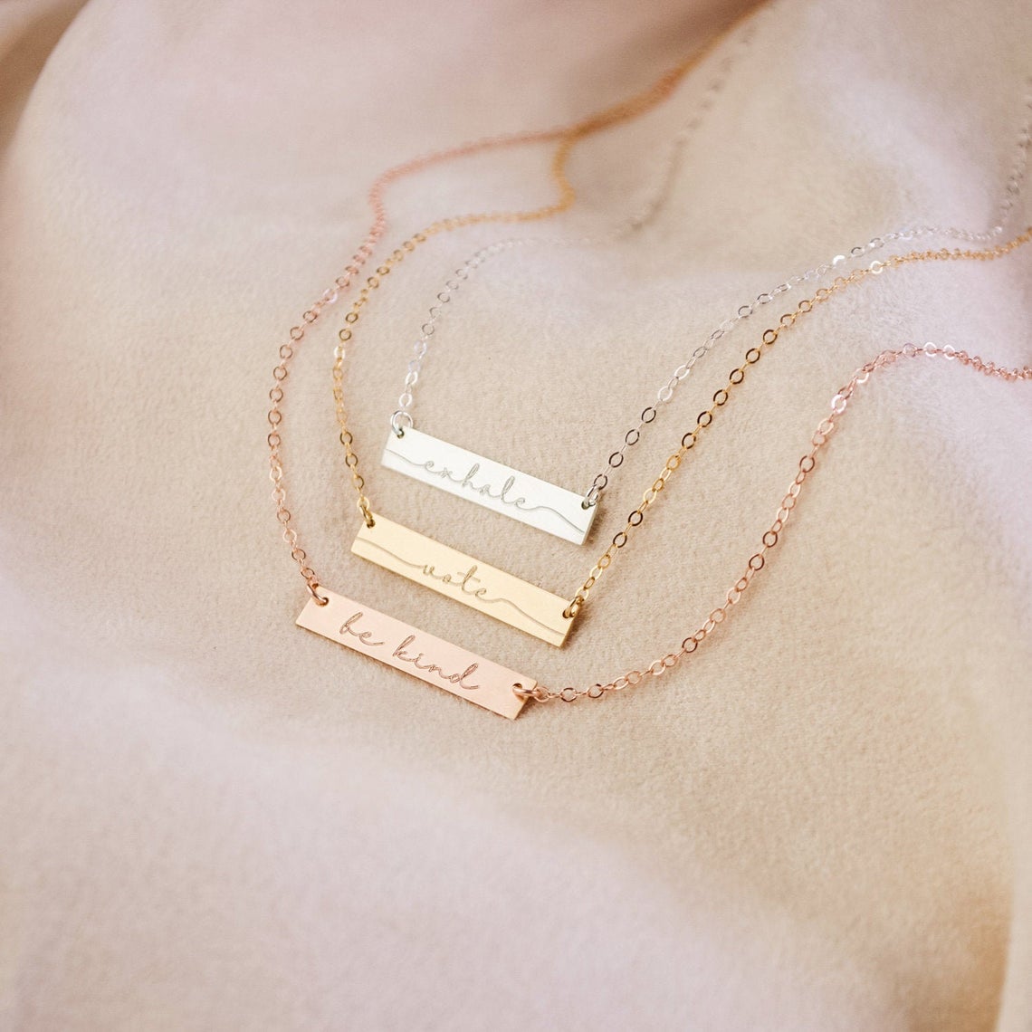 a silver, gold and rose gold necklace on a peach background. Personalized bar necklace for special events Custom bar pendant with initials Engraved bar necklace for her special occasion Personalized minimalist bar necklace with message Custom bar necklace for personalized moments Engraved bar pendant for holidays Personalized bar necklace for unique gifts Custom minimalist bar jewelry piece