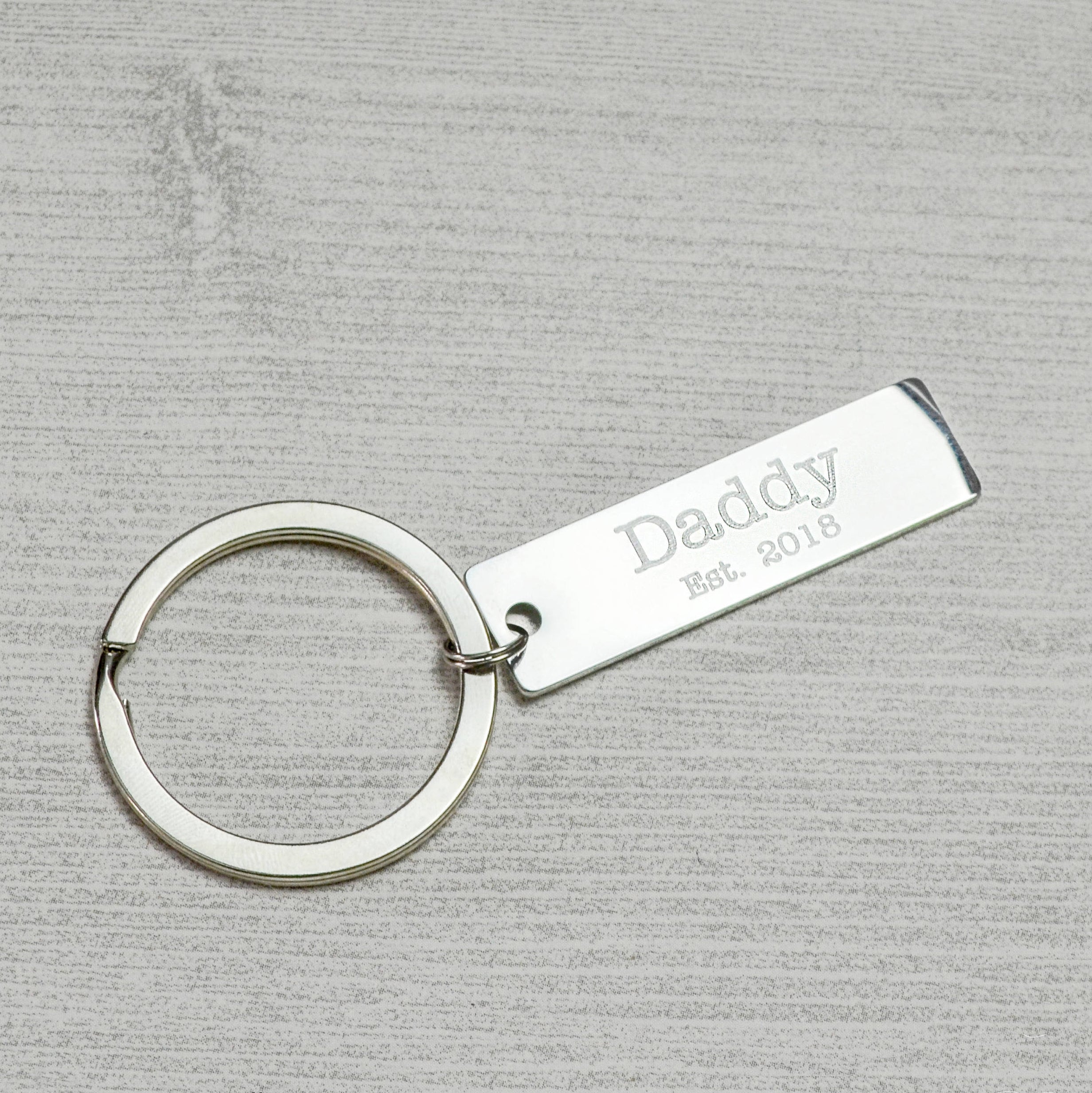 A silver engraving keychain with Daddy Est 2018 in Typewriter font on the back of a heartbeat keychain. The keychain sits on a gray background. What better gift for a new parent than their babys first heartbeat forever preserved in an elegantly simple keychain?Silver Metal Keychain Personalized Gift for New Parent 