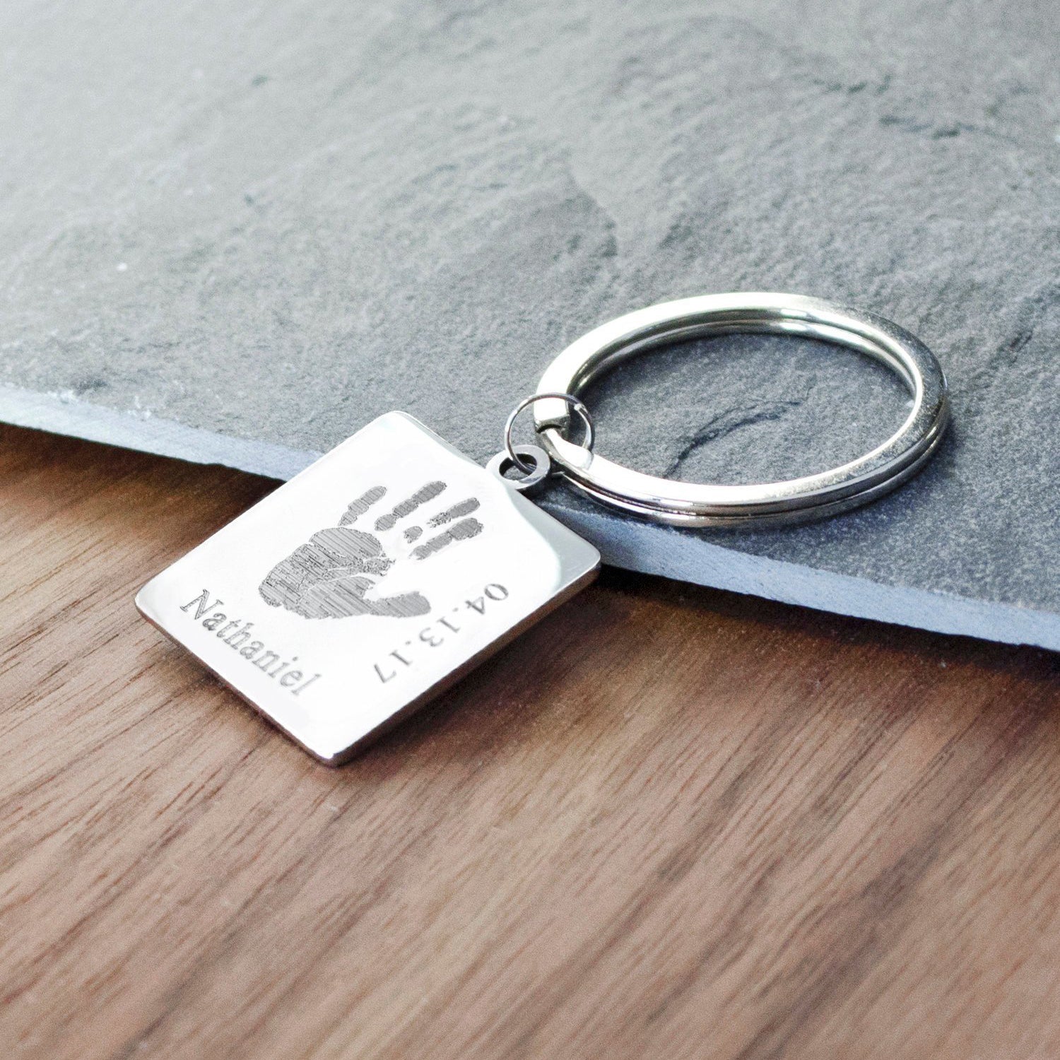 Square Stainless Steel Keychain Custom Engraved with Baby Handprint, Name, and Birth Date Resting on a gray stone tile and wood. 